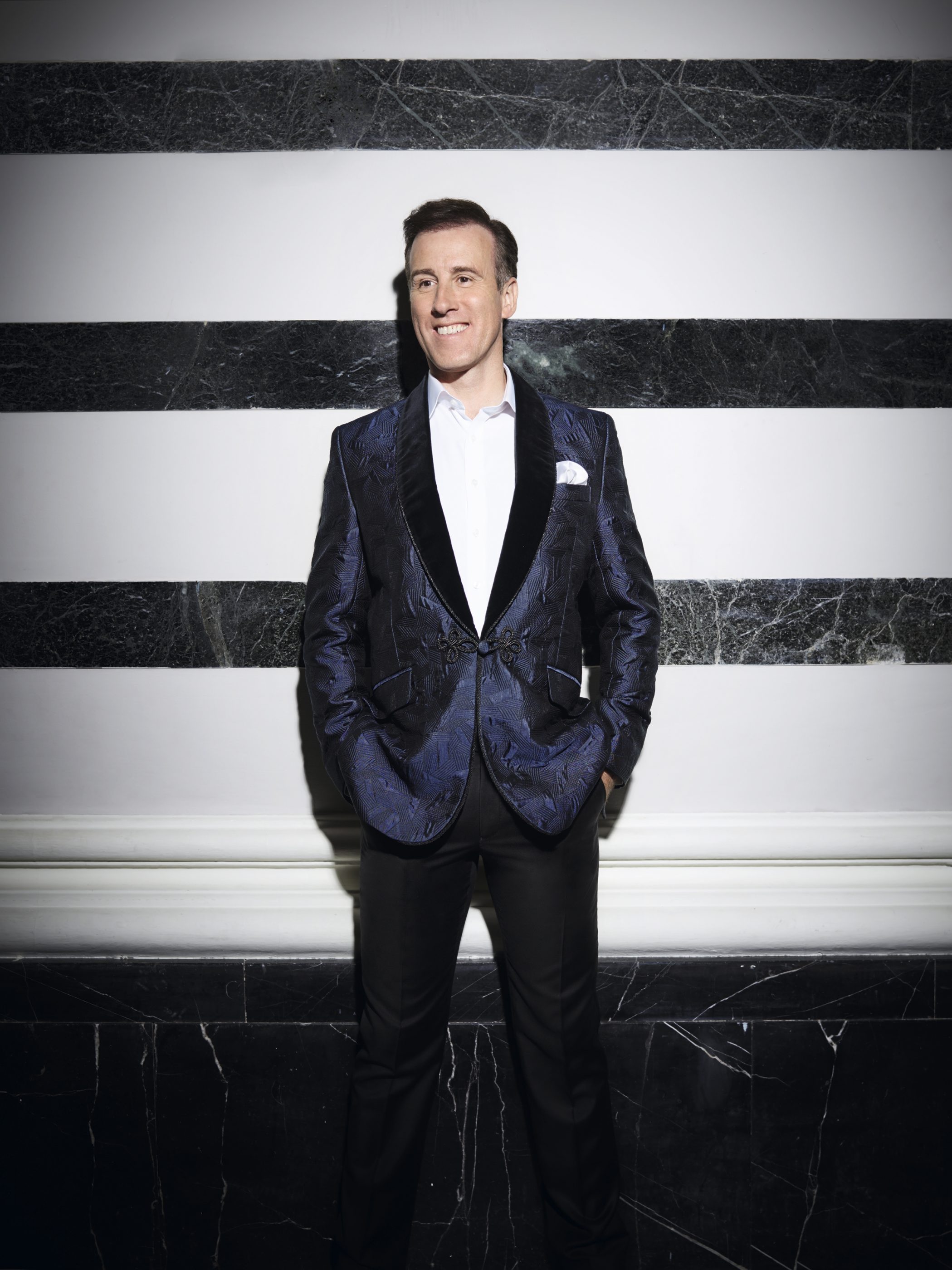 Du Beke in a shiny suit jacket, stood in front of a black and white striped wall.
