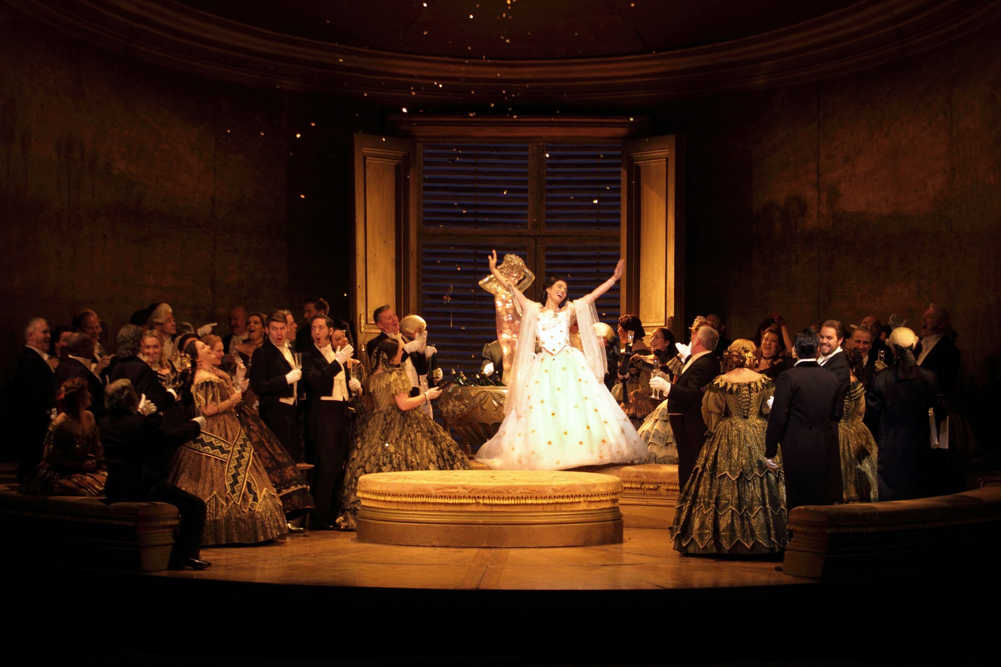 An opera singer in a white ball gown being cheered by the ensemble surrounding her.