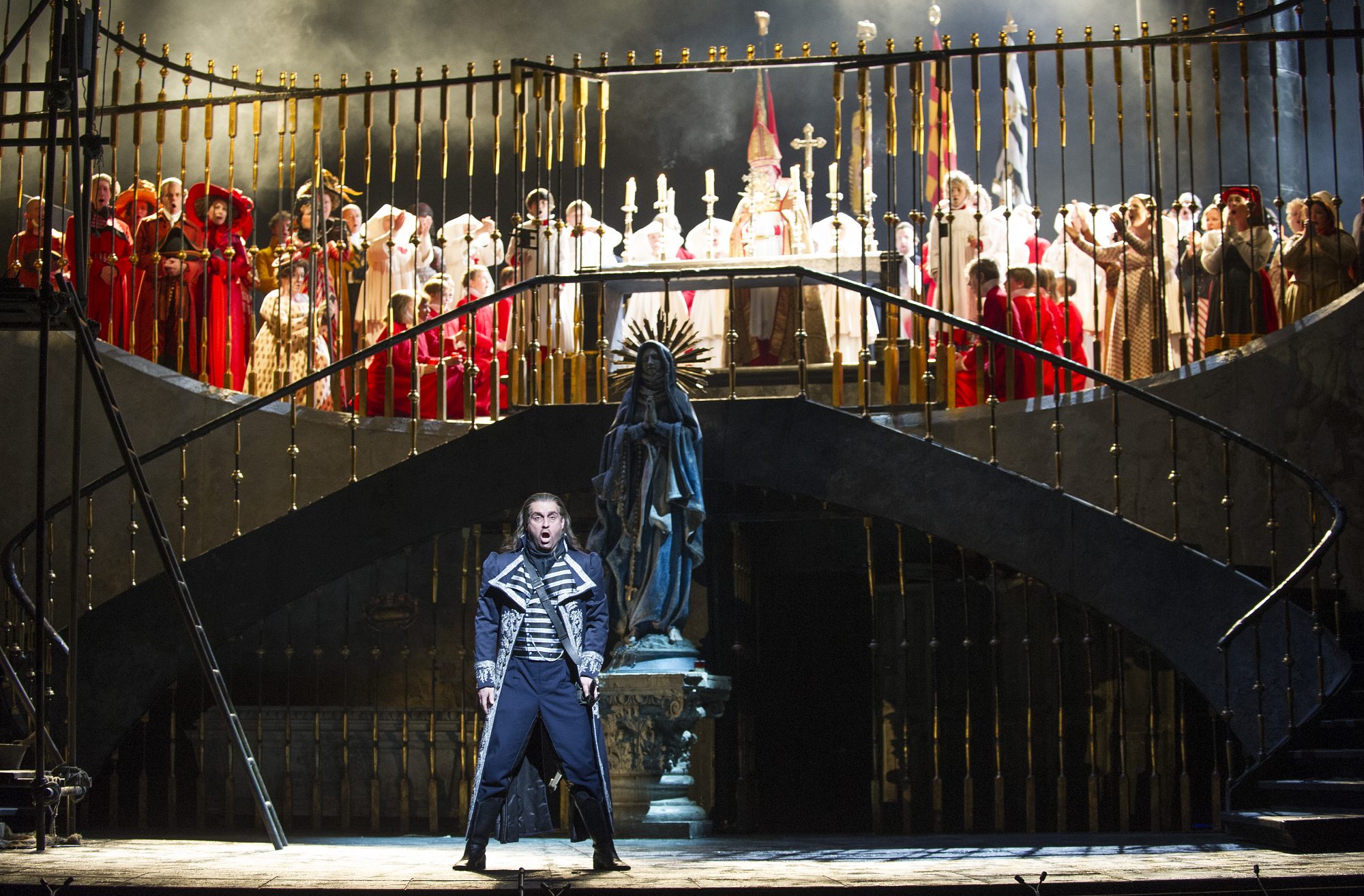 An opera singer singing on stage, with the chorus dressed in red robes stood on a balcony above him.