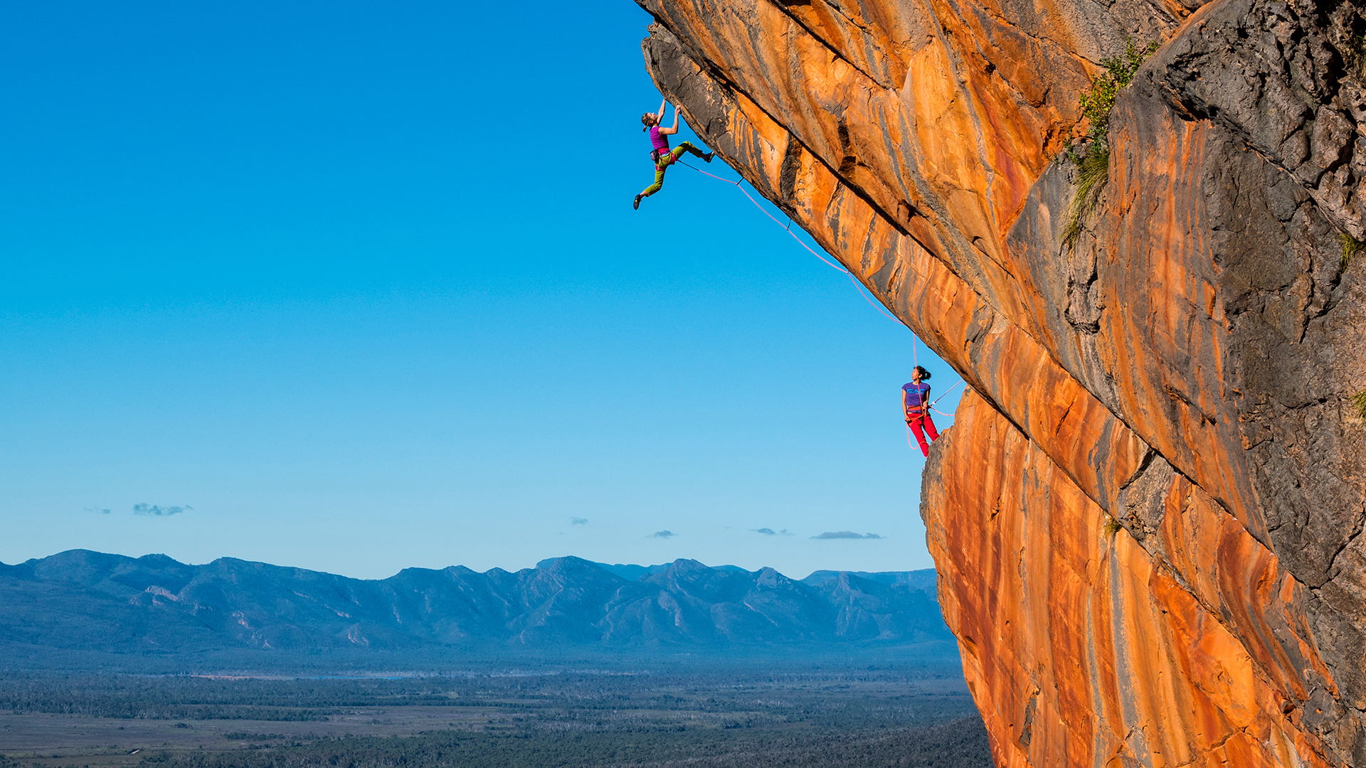 2 climbers scaling an orange/brown rock face. A clear blue sky and mountain range in the distance.