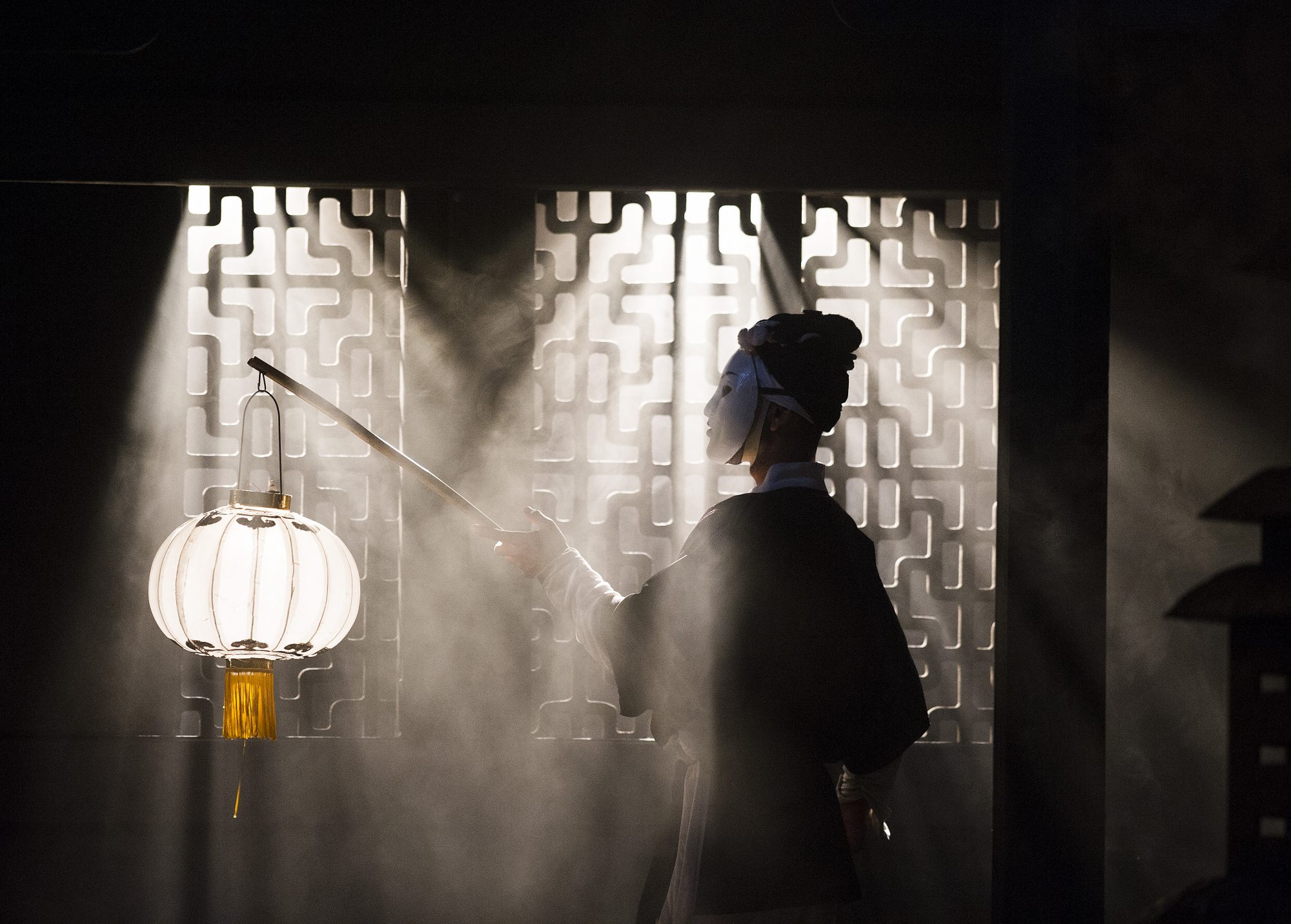 A person with a white mask covering their face, holding a lantern.