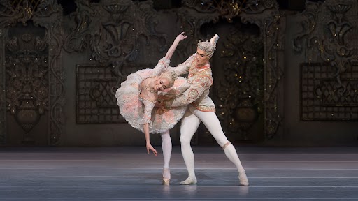 Two ballerinas (man and woman) dance together as the man holds the woman as she leans.