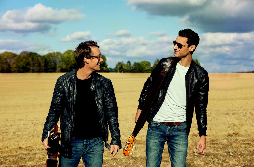 The band duo dressed in leather jackets and holding guitars walking through a field