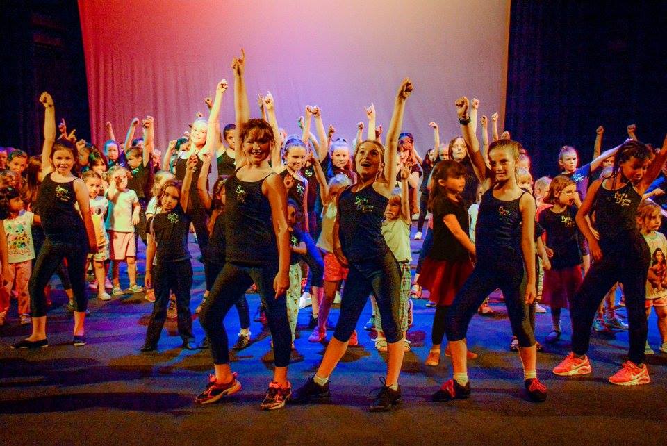 A variety of dancers with arms in the air, on a colourfully lit stage