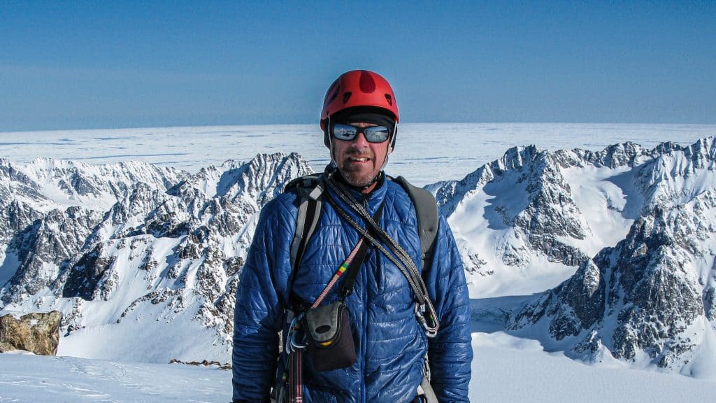 Photo shows Simon Yates in mountaineering gear at the peak of a snow capped mountain.
