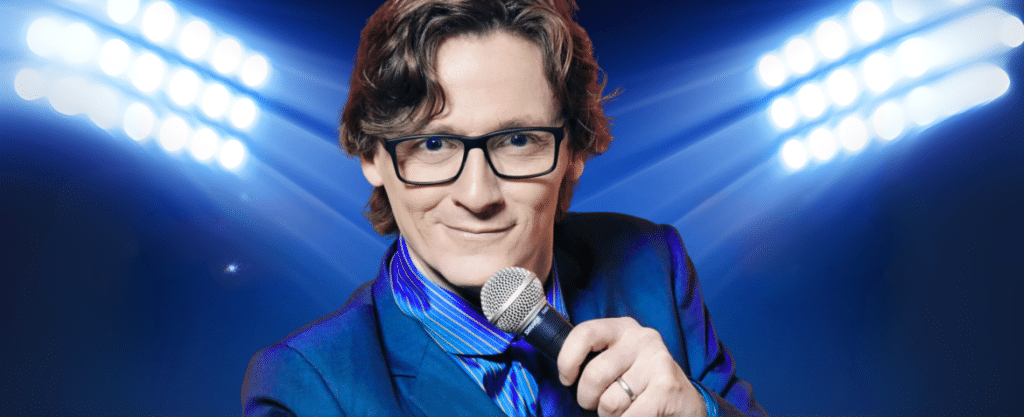 Ed Byrne wearing black glasses, wearing a blue suit and holding a microphone