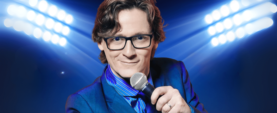 Ed Byrne wearing black glasses, wearing a blue suit and holding a microphone